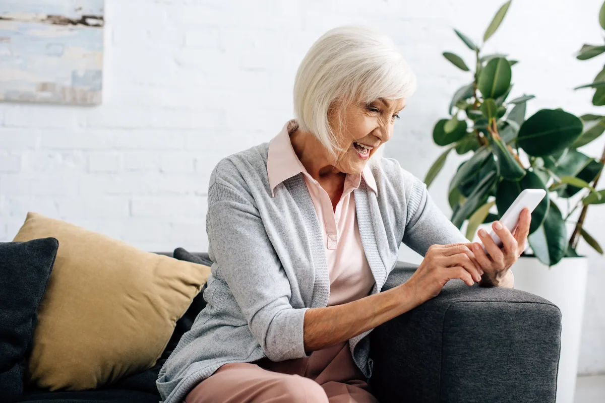 Ältere Frau auf dem Sofa, lacht beim Blick auf ihr Smartphone